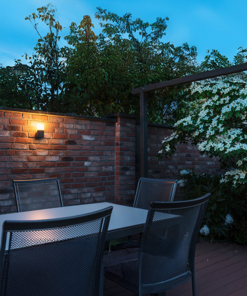 Mediterrane Natursteinmauer mit warmweiß leuchtender Außenwandlampe hinter einer gemütlichen Gartentischgruppe